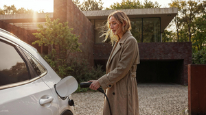 Vrouw in beige jas laadt haar witte wagen op voor een moderne villa met cortenstalen wand